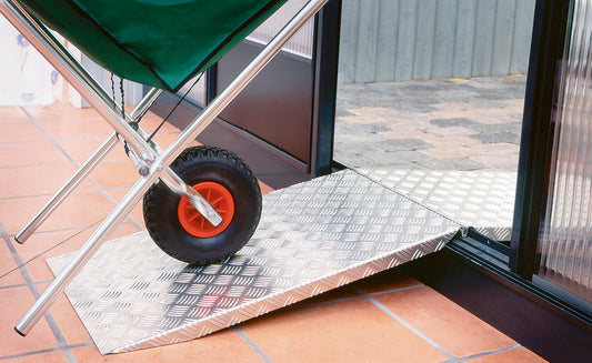 Auffahrrampe aus geriffeltem Aluminium an der Türschwelle eines Gewächshauses, darüber rollt das Rad einer Schubkarre